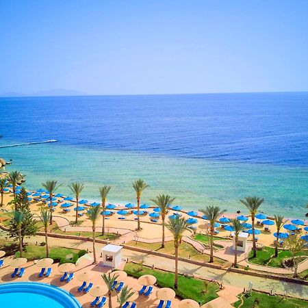 Pickalbatros Royal Grand Sharm - Adults Friendly 16 Years Plus Sharm El Sheikk Exterior foto The photo displays a beautiful coastal scene featuring a clear blue sea that transitions into green tones near the shore. In the foreground, there are sandy beaches lined with palm trees and a variety of beach umbrellas. There is also a swimming pool