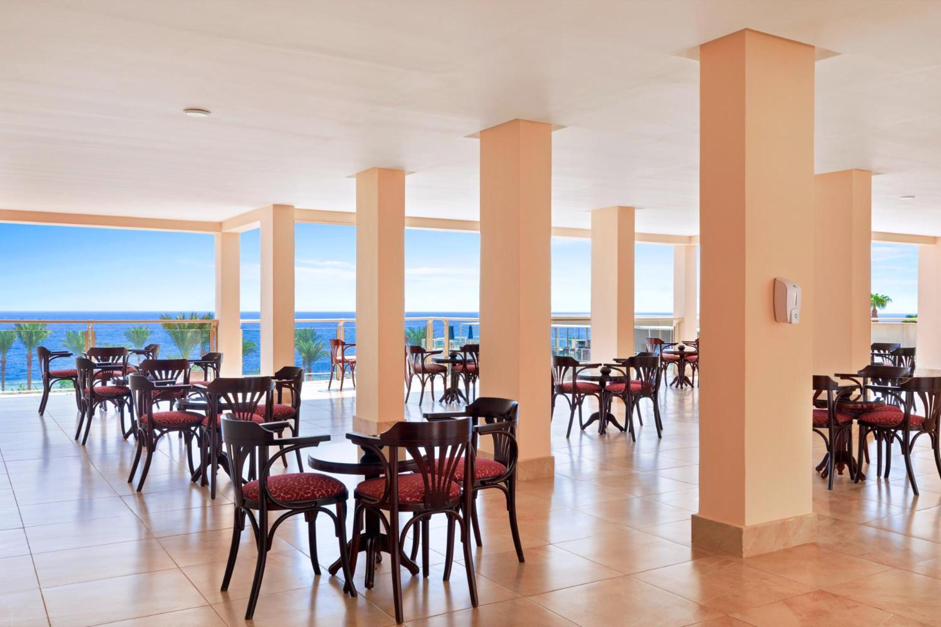 Pickalbatros Royal Grand Sharm - Adults Friendly 16 Years Plus Sharm El Sheikk Exterior foto The image shows a dining area or café with several round wooden tables and chairs, featuring red upholstery. The space is well-lit, with large windows that allow a view of the ocean in the background, contributing to a bright and open atmosphere. The