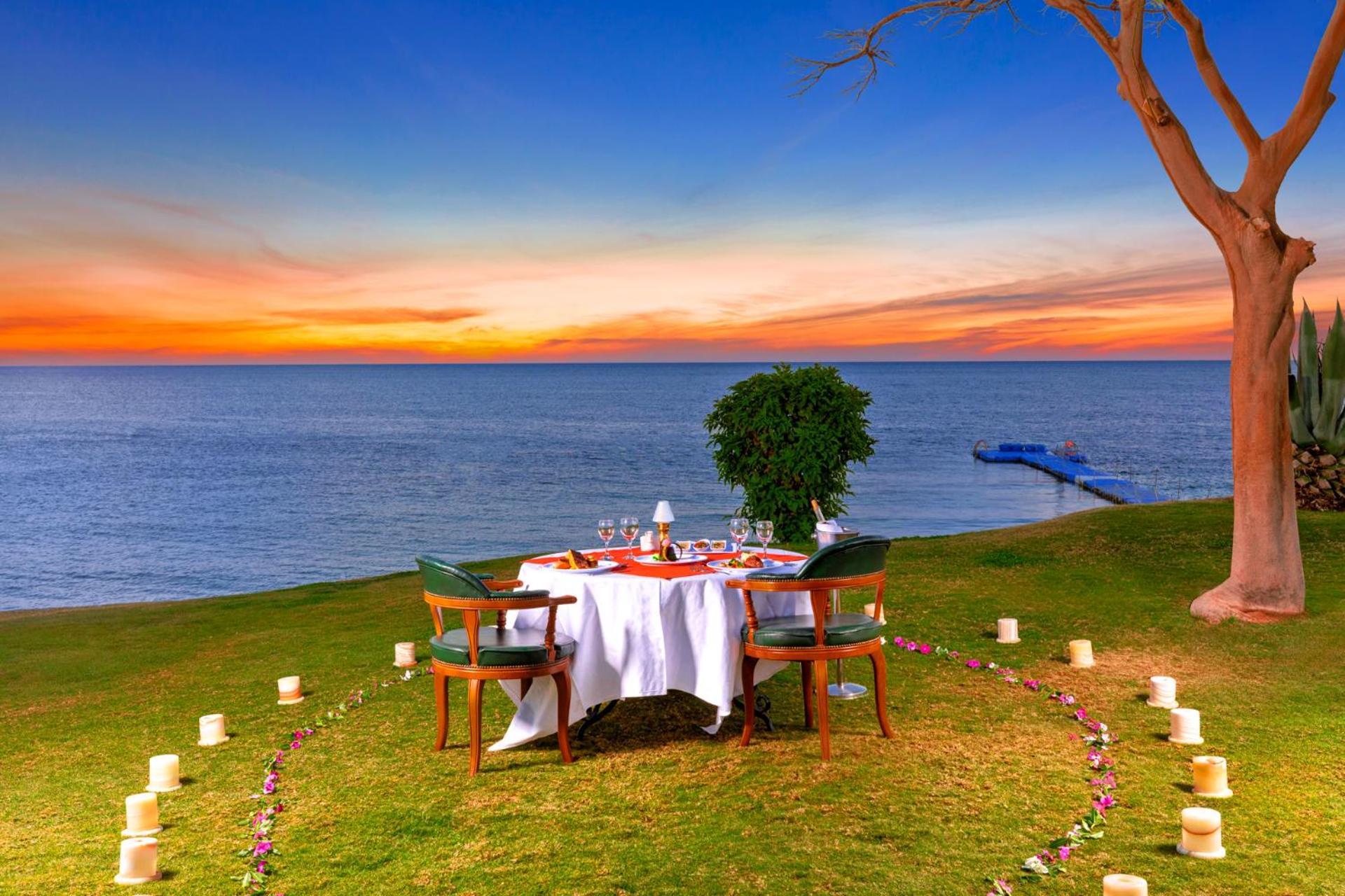 Pickalbatros Royal Grand Sharm - Adults Friendly 16 Years Plus Sharm El Sheikk Exterior foto The photo depicts a romantic outdoor dining setup by the seaside. In the foreground, there is a round table elegantly set with a white tablecloth, plates, and glassware, creating a cozy atmosphere. Surrounding the table are green chairs. A circle of 