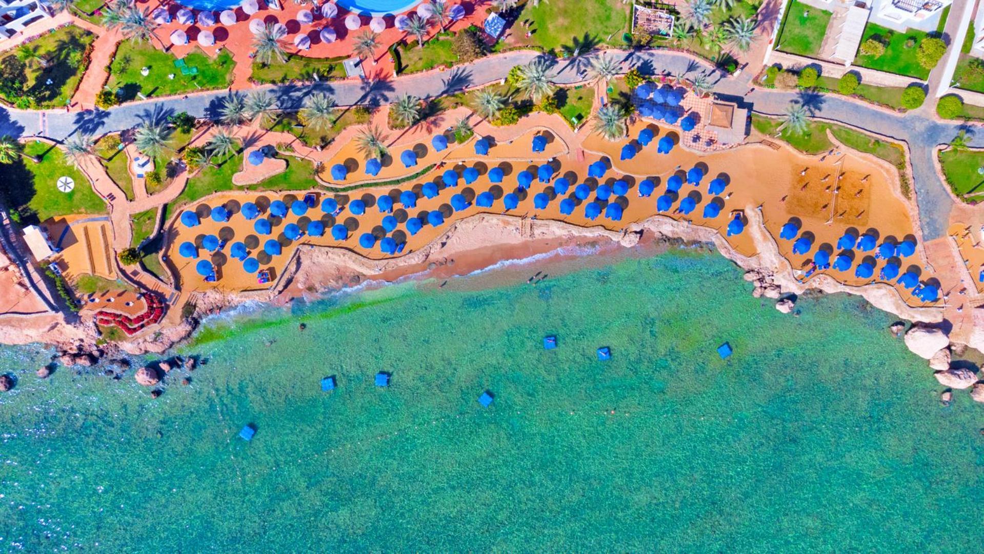 Pickalbatros Royal Grand Sharm - Adults Friendly 16 Years Plus Sharm El Sheikk Exterior foto The photo shows an aerial view of a beach resort area. You can see a stretch of golden sandy beach lined with numerous blue umbrellas. The beach is bordered by clear turquoise waters. There are palm trees scattered throughout the area, adding to the 