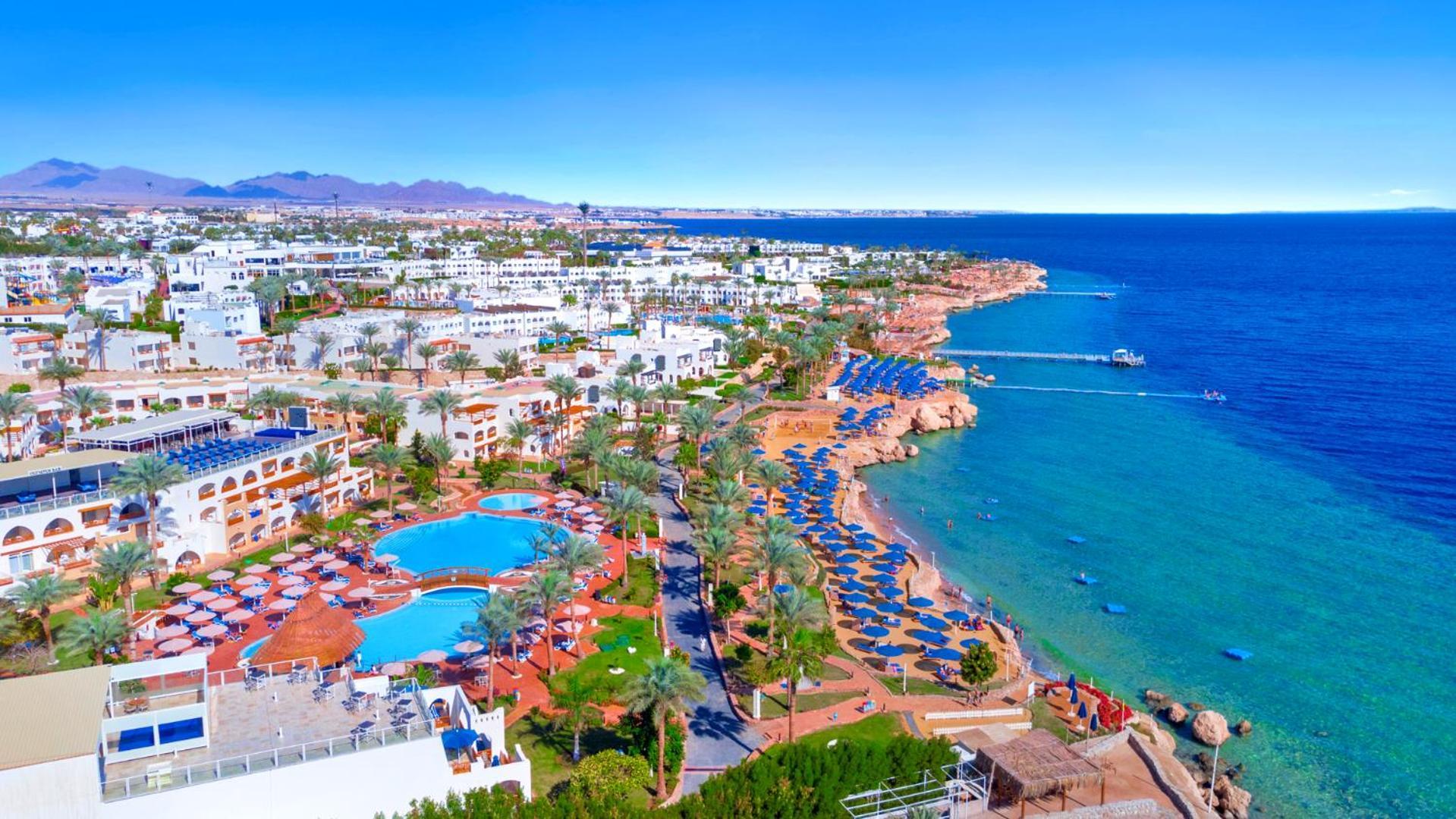 Pickalbatros Royal Grand Sharm - Adults Friendly 16 Years Plus Sharm El Sheikk Exterior foto The photo features a picturesque coastal scene, likely from a resort area. In the foreground, there is a large swimming pool surrounded by lounge chairs. Further back, the landscape includes several buildings, possibly hotels or apartments, that over