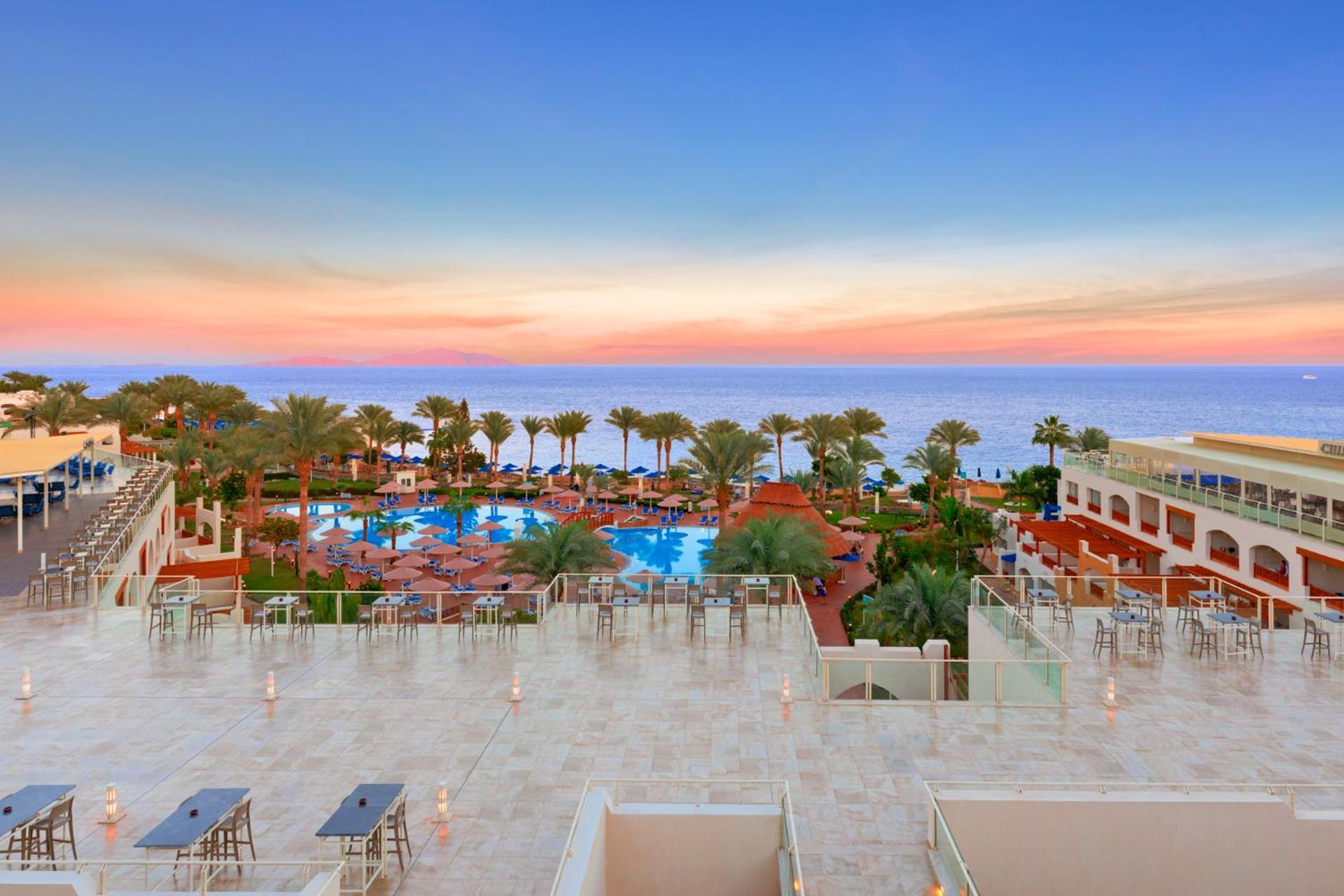 Pickalbatros Royal Grand Sharm - Adults Friendly 16 Years Plus Sharm El Sheikk Exterior foto The photo depicts a beautiful resort scene at sunset. In the foreground, there is a spacious terrace area with modern furniture. Below, a large swimming pool is surrounded by lush palm trees and lounge chairs, indicating a relaxing atmosphere. The vi