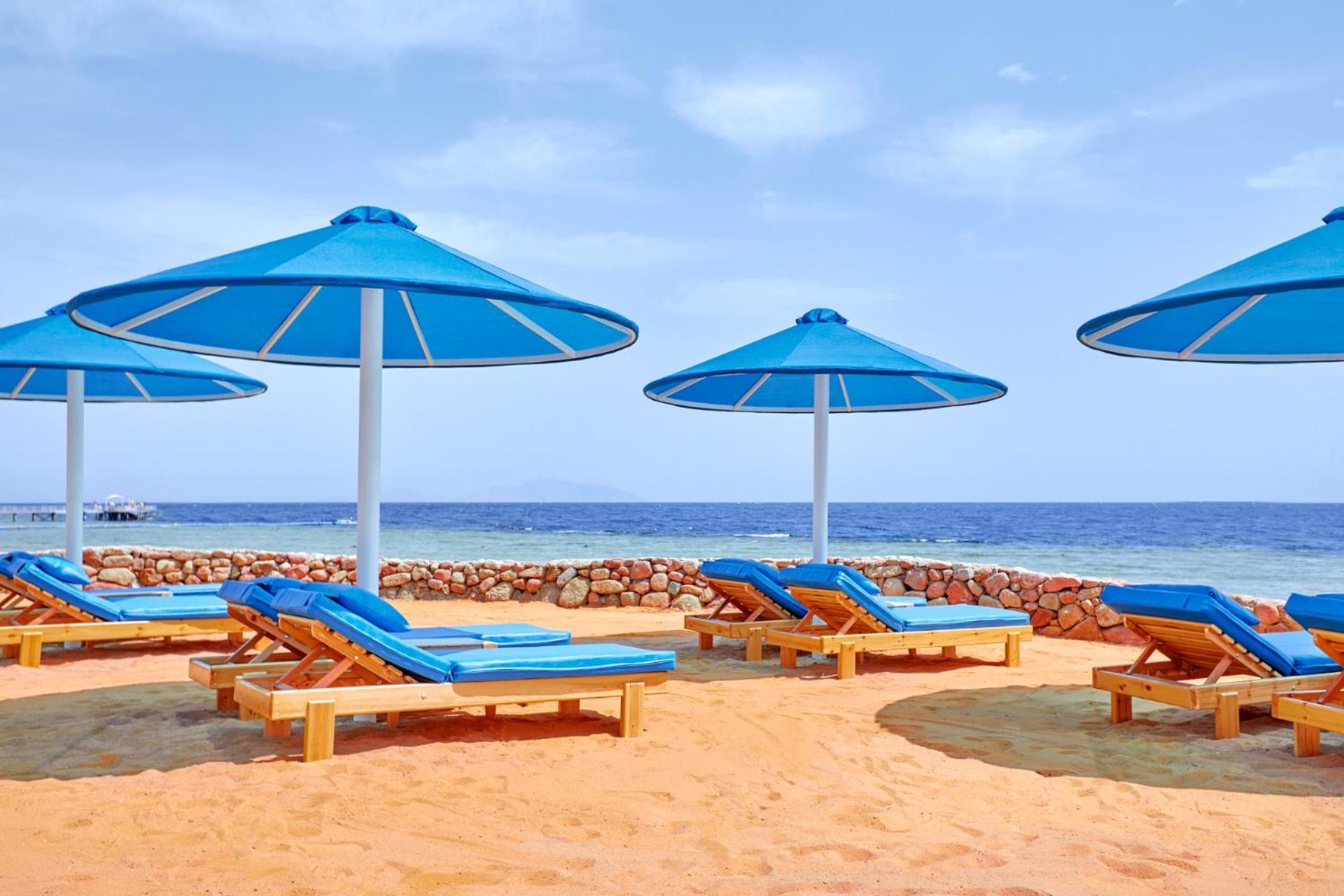 Pickalbatros Royal Grand Sharm - Adults Friendly 16 Years Plus Sharm El Sheikk Exterior foto The photo shows a beach scene featuring several blue umbrellas providing shade over wooden lounge chairs. The sandy beach extends toward a calm sea, creating a relaxing atmosphere. The sky is mostly clear, with a few clouds, suggesting pleasant weath