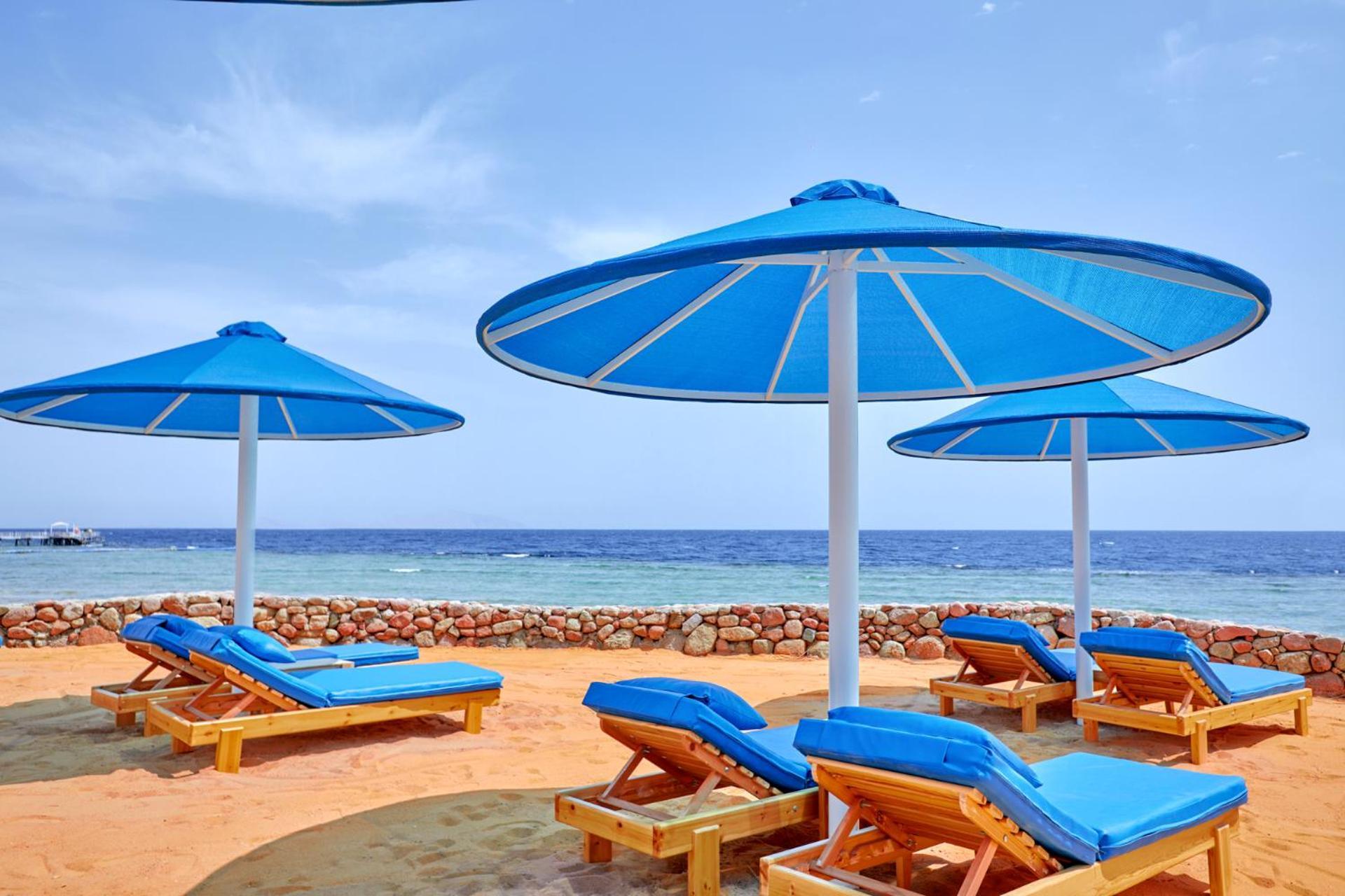 Pickalbatros Royal Grand Sharm - Adults Friendly 16 Years Plus Sharm El Sheikk Exterior foto The photo shows a serene beach scene with several blue sun loungers arranged beneath large blue umbrellas. The loungers are positioned on sandy ground, with the ocean visible in the background. The sky is clear, indicating a bright and sunny day, and