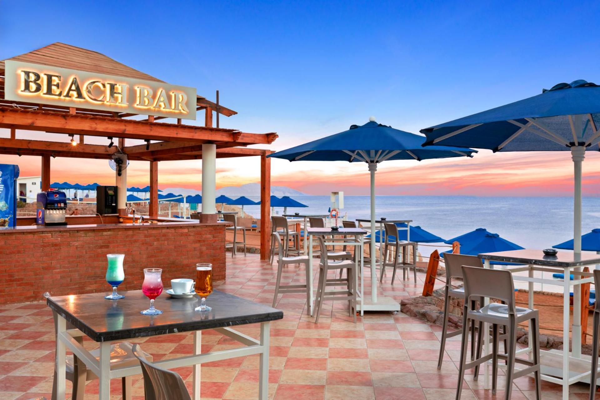 Pickalbatros Royal Grand Sharm - Adults Friendly 16 Years Plus Sharm El Sheikk Exterior foto The photo shows a beach bar setting, featuring a wooden structure with the words "BEACH BAR" prominently displayed. There are several tables and chairs arranged on a tiled floor, with some drinks placed on the tables. Blue umbrellas provide shade for